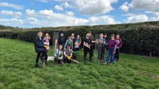 A group of people stand in a field with newly planted trees, holding spades and a wheelbarrow