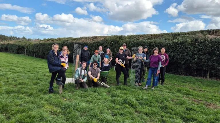 A group of people stand in a field with newly planted trees, holding spades and a wheelbarrow