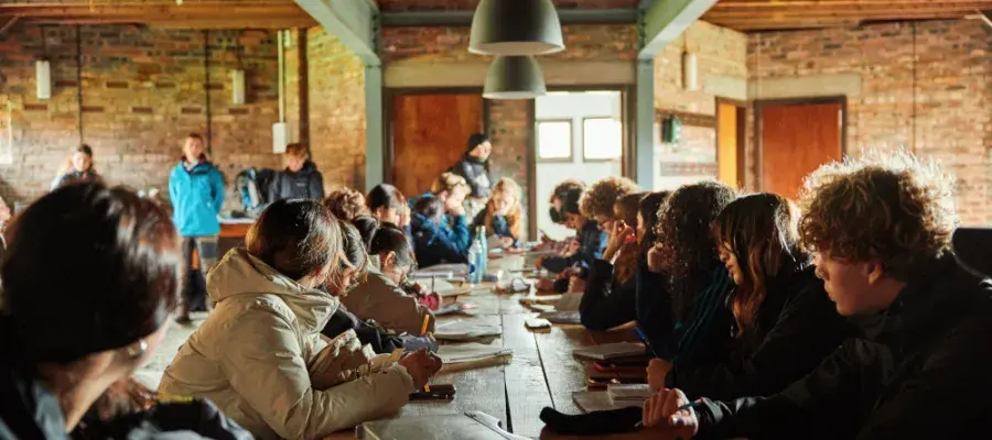 People sitting at a long table with notebooks listening to a speaker.