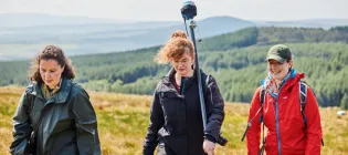 Three people walk across a hillside with rucksacks and technical equipment