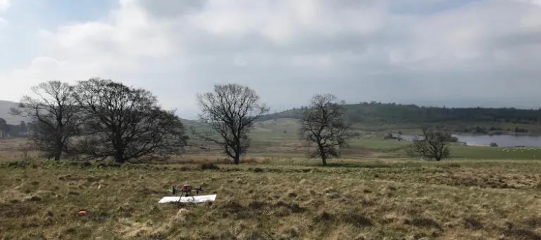 Drone sitting on a drone pad in a field of grass with a line of trees in the background, and a blue sky with some clouds.