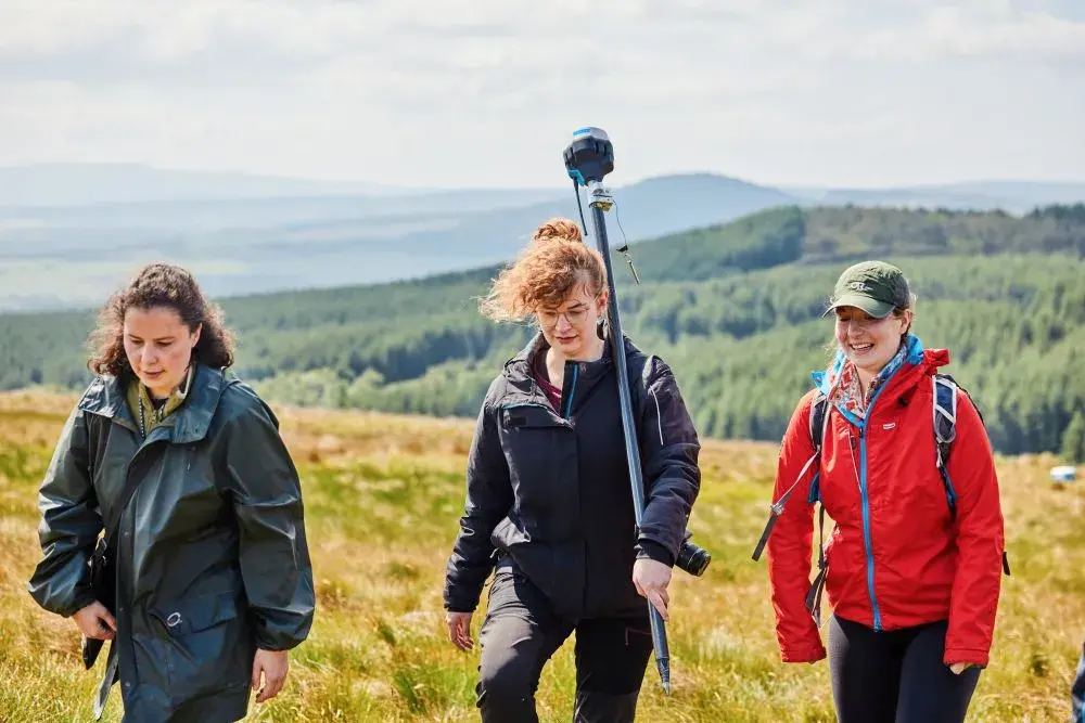 Three people walk across a hillside with rucksacks and technical equipment