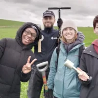 four people stand in a field and smile at the camera holding fieldwork tools