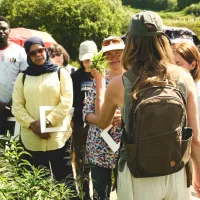A woman speaks to a group of people, with leaves of a plant in her hand. Trees are visible in the background.