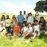 A large group of people on a sunny day in a grassy area with trees providing some shelter and water visible in the left.