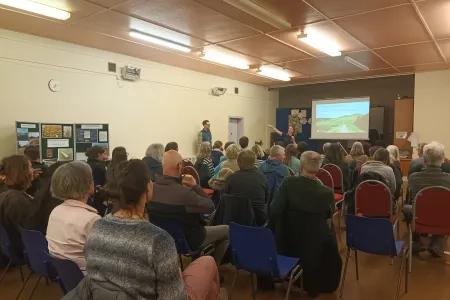 A photgraphy of an audience of people from behind in a community hall, listening to a presentation from Julie Wilson, Community Ranger