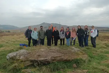 A group of people gathered round a fallen standing standing stone in a grassy field.