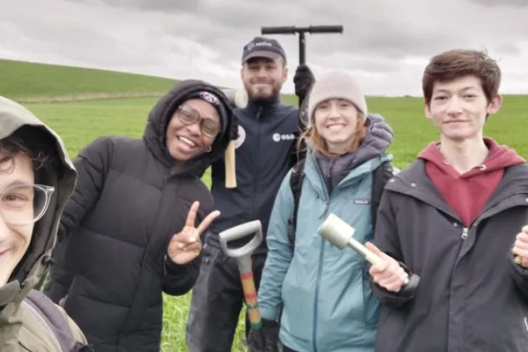 four people stand in a field and smile at the camera holding fieldwork tools