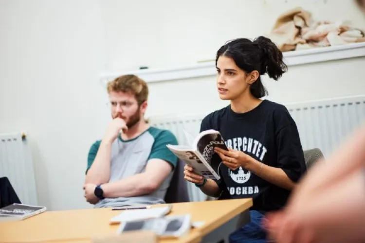 Two people sit at a desk, with one holding a book