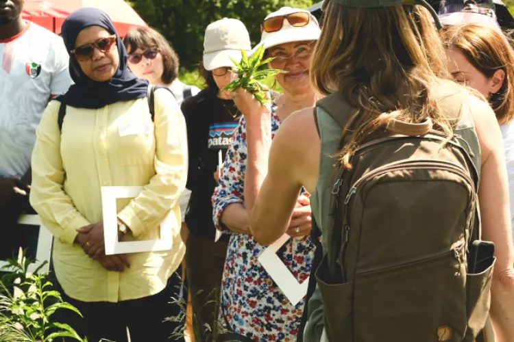 A woman speaks to a group of people, with leaves of a plant in her hand. Trees are visible in the background.