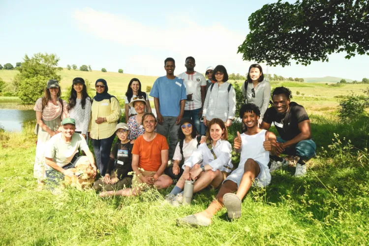 A large group of people on a sunny day in a grassy area with trees providing some shelter and water visible in the left.