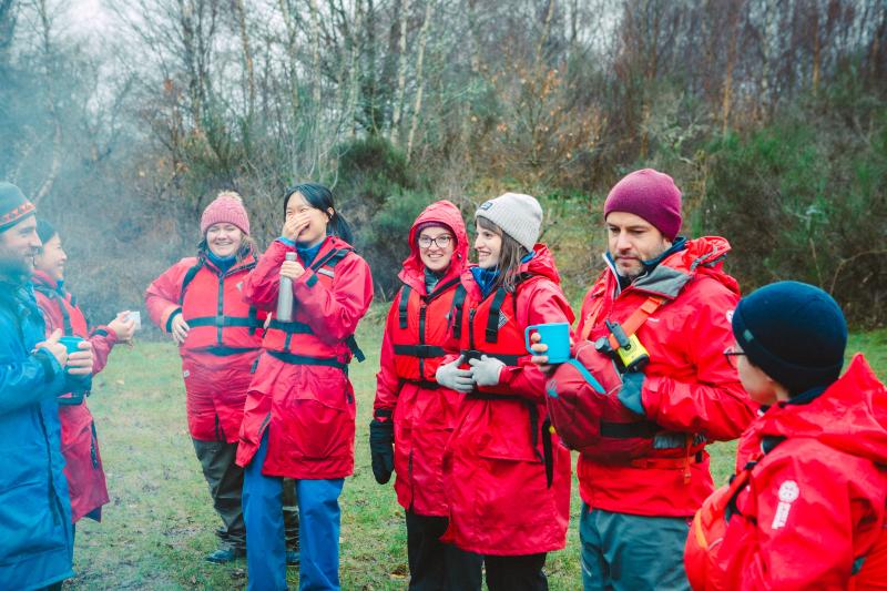 Students and staff wearing red life jackets and coats stand outside laughing and smiling