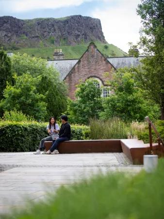 two people sit on a bench surrounded by vegetation with crags in the background