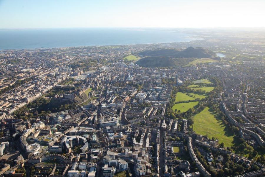 An aerial photograph of central Edinburgh showing the meadows and Holyrood Park in the background