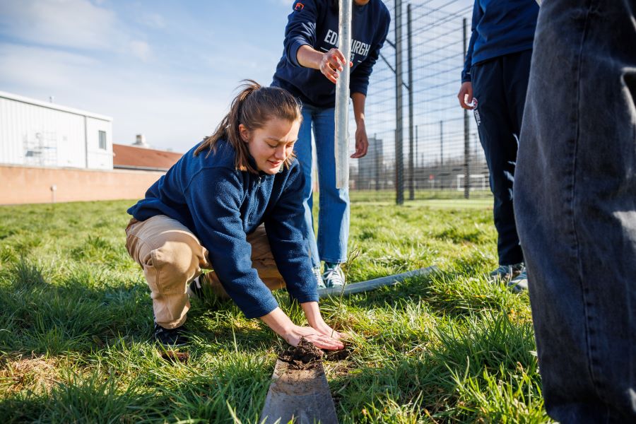 A student athlete plants a sapling at Peffermill Playing Fields, helped by other students